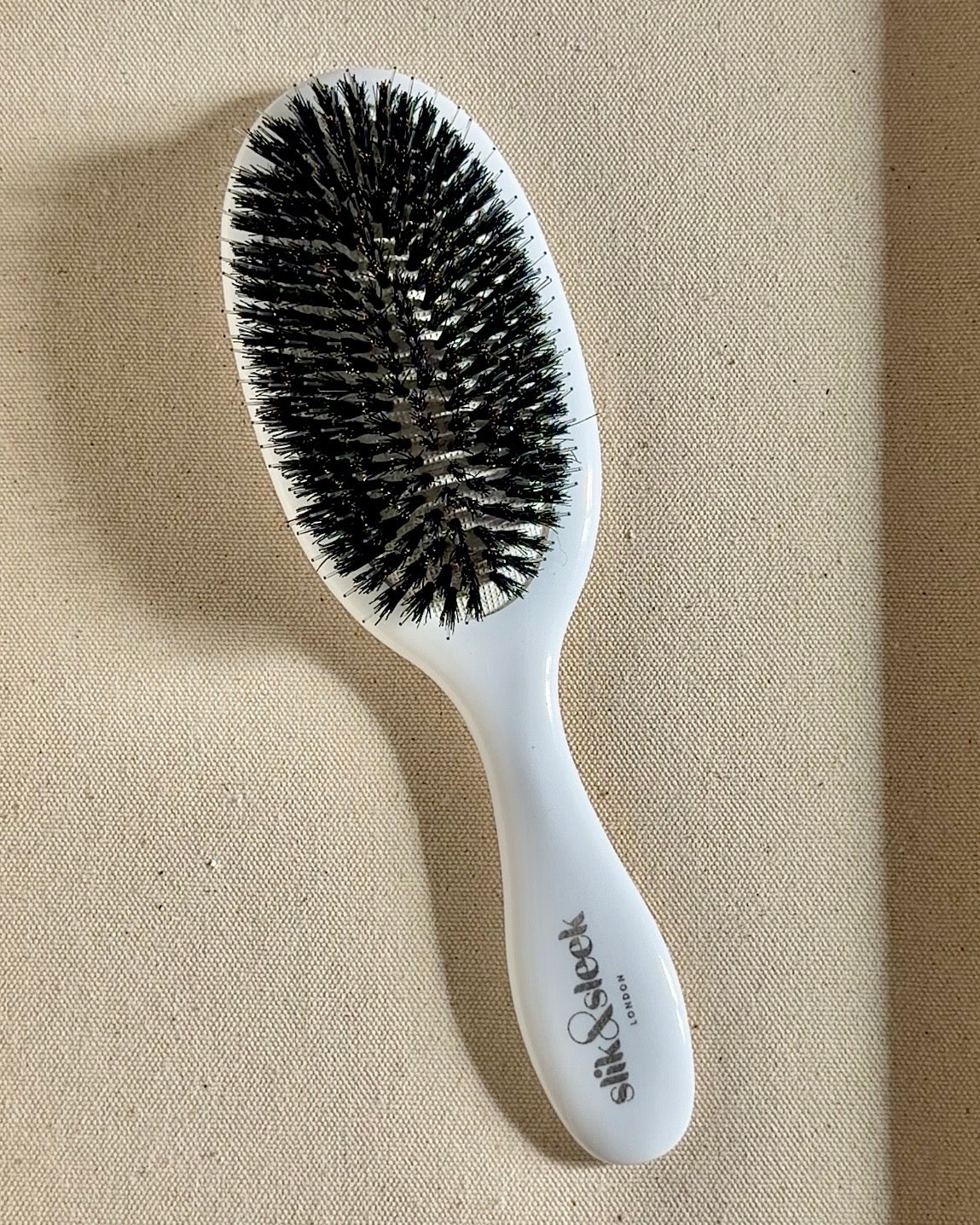 White hairbrush with black mixed boar and nylon bristles on a beige fabric background - Slik & Sleek London salon quality brush