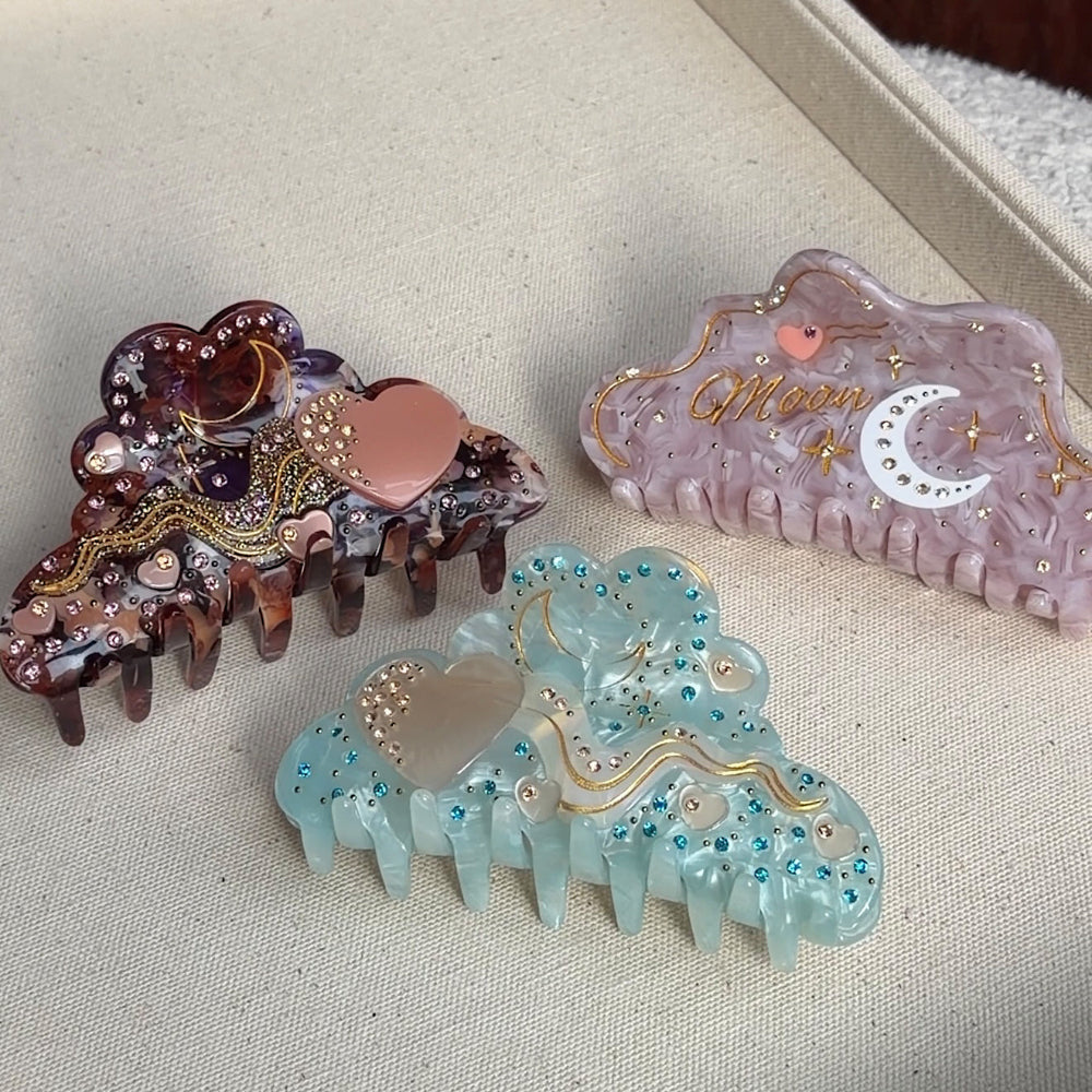 A trio of 10cm celestial-themed acetate hair clips in pink with a moon motif, burgundy with a heart, and marbled teal with rhinestone accent