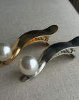 Two pearl hair clips, one gold and one silver, on a textured surface.